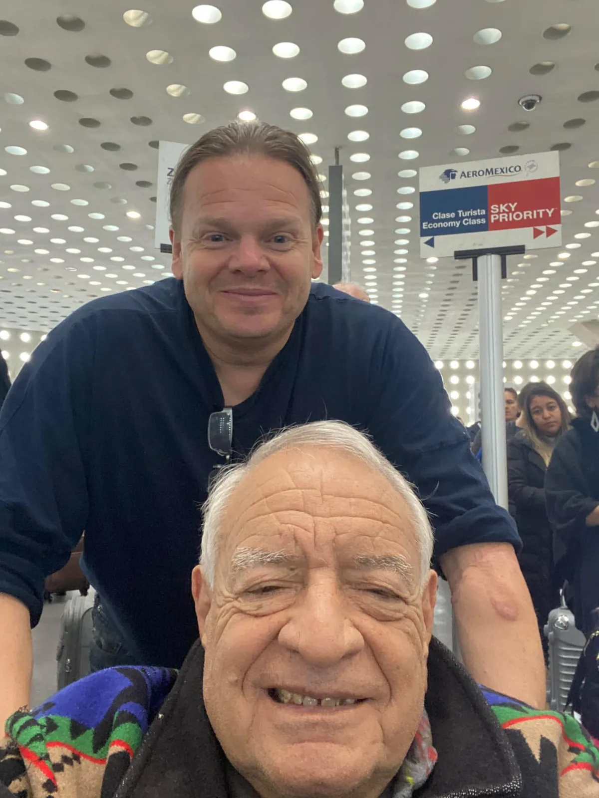 Selfie of two smiling men in an Aeroméxico airport terminal: an elder with white hair in a patterned blanket, and a younger man in a navy shirt leaning over him from behind.