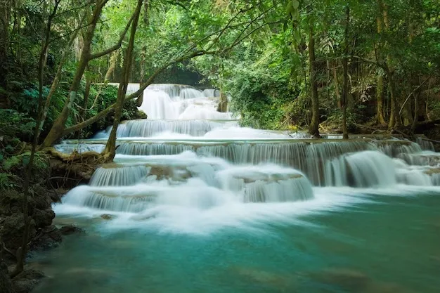 A multi-tiered waterfall cascading over limestone steps into a turquoise pool, surrounded by lush green forest.