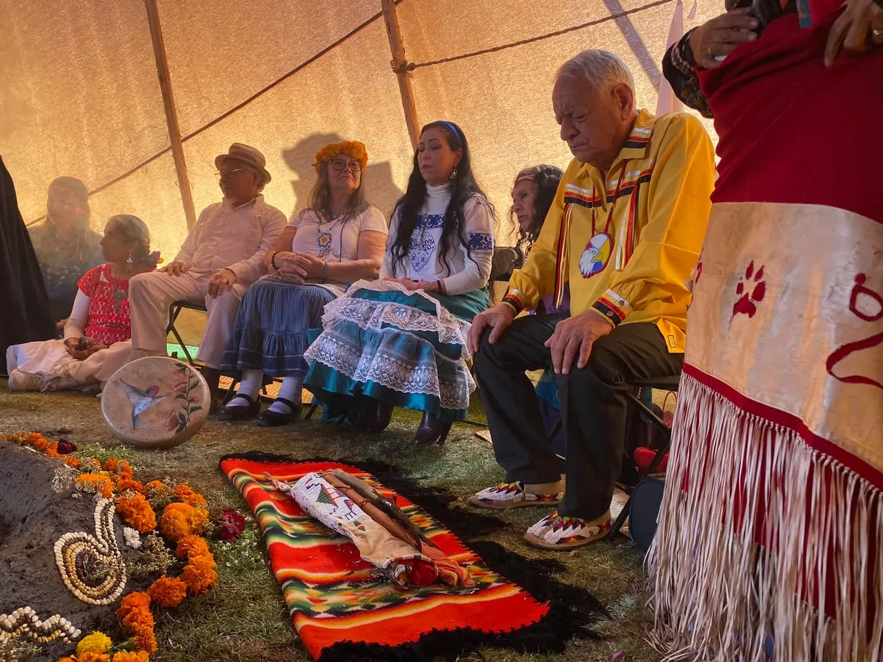 Elders and participants in traditional clothing sit in prayer inside a large tipi, near a sacred bundle on a striped red blanket, marigolds and a hummingbird-painted drum.