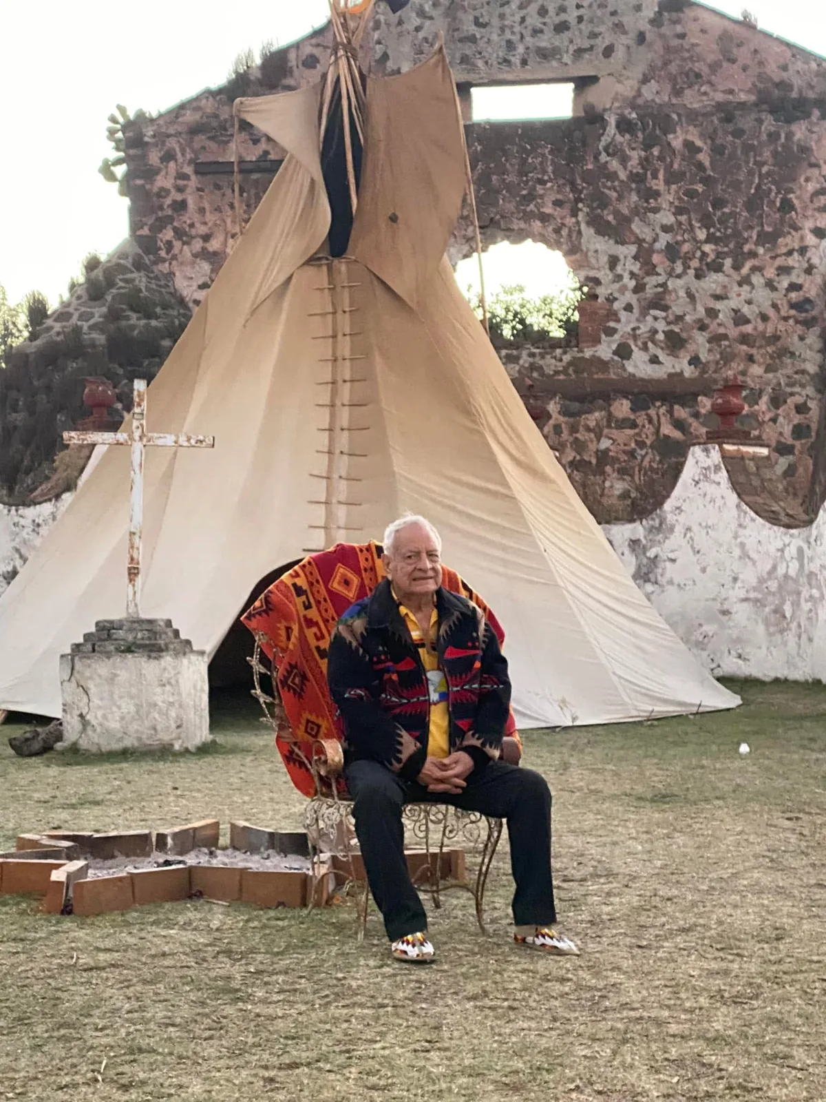 An elderly man with white hair, in a patterned jacket and beaded moccasins, sits on an ornate chair draped with a red blanket before a tipi, a white cross and old stone ruins.