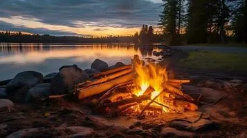 A campfire of stacked logs burns among rocks on the shore of a calm lake at sunset, with dark evergreen trees silhouetted against a golden sky.