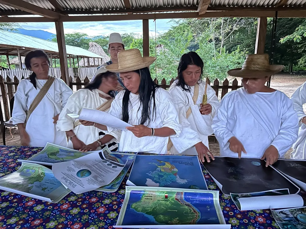 People in white traditional clothing, some in straw hats, stand under an open shelter studying printed bioregional maps of the Americas spread on a flowered tablecloth.