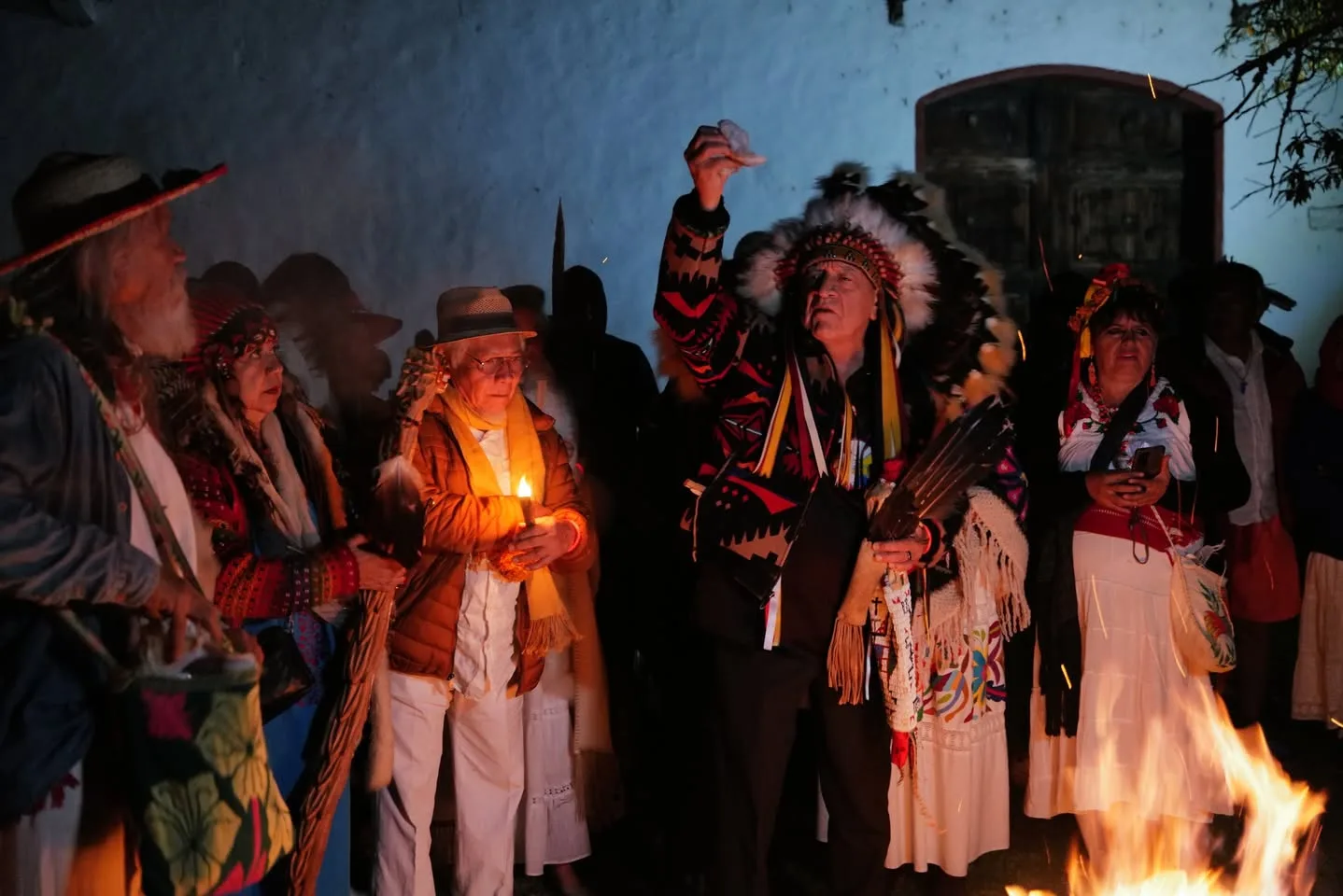 At night beside a fire, an elder in a feather headdress raises his hand in prayer, surrounded by people in traditional dress, one holding a candle.