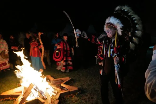 An elder in a feather headdress and patterned jacket stands by a blazing fire at night as people in traditional clothing gather behind him.