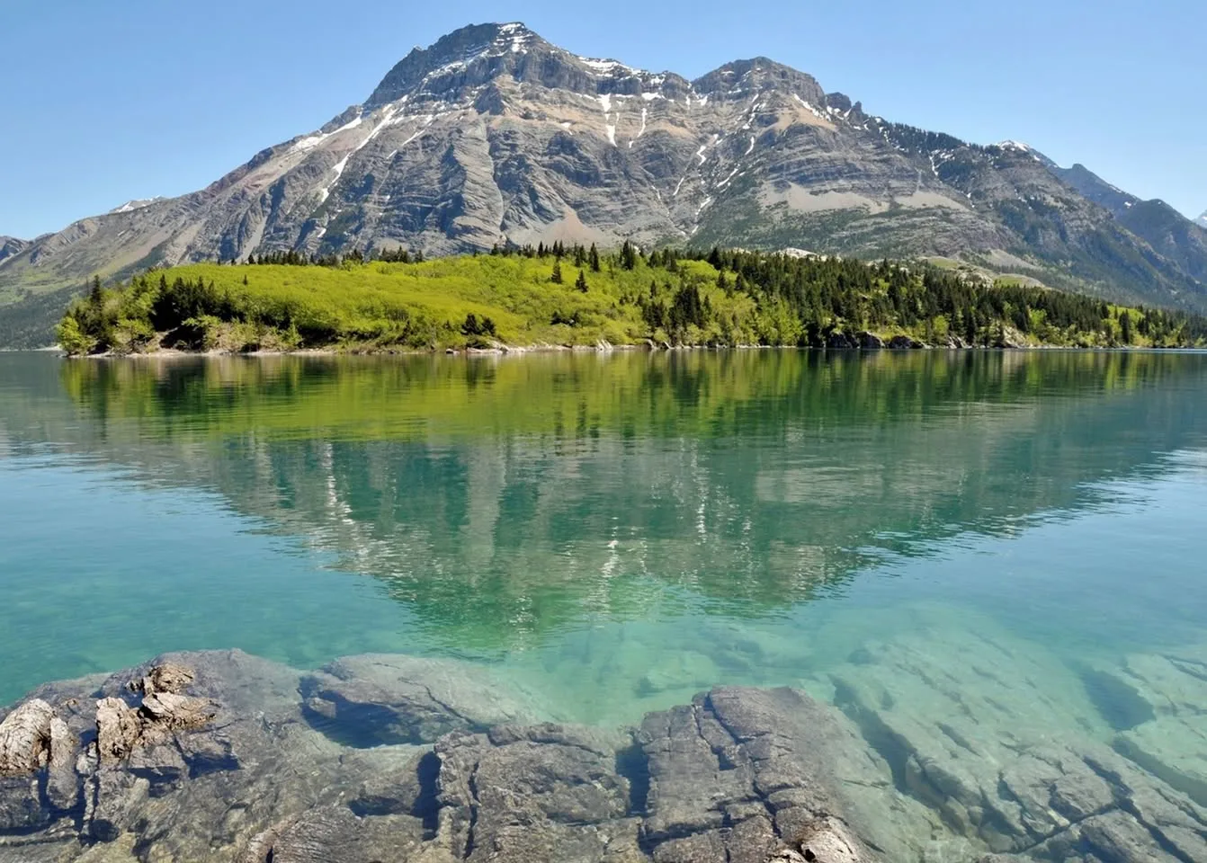 A snow-streaked mountain rises behind a green forested shoreline, reflected in a calm, clear turquoise lake with rocks visible beneath the water in the foreground.