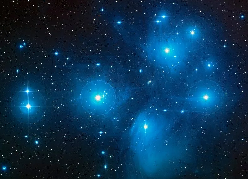 The Pleiades star cluster, several bright blue-white stars glowing within wisps of blue nebula against a dark sky scattered with smaller stars.