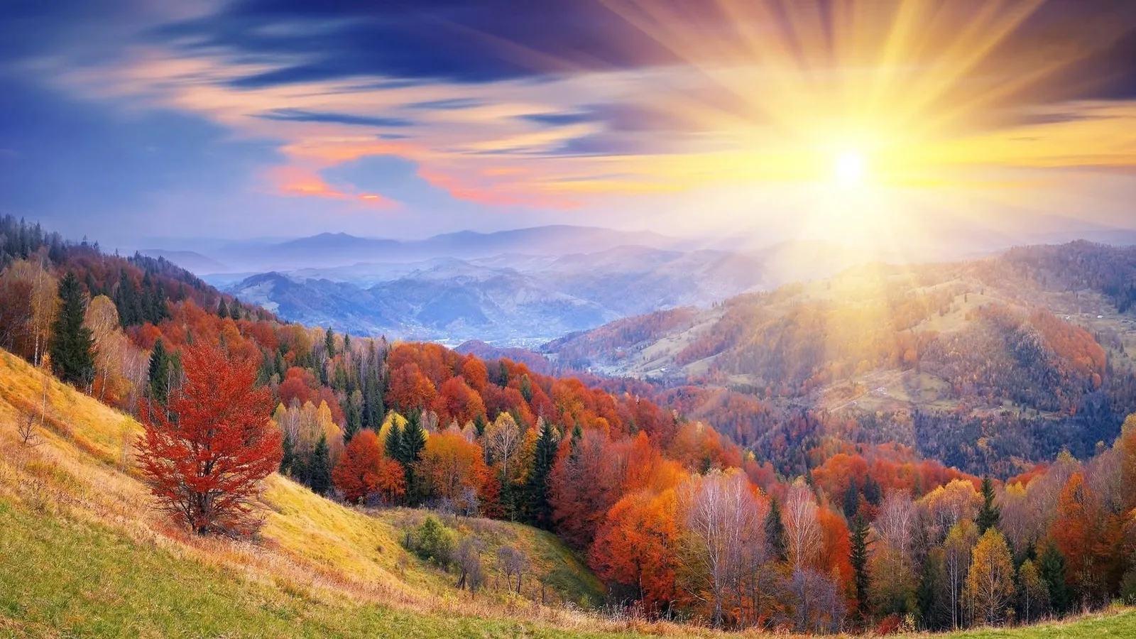Sunlight bursts over misty mountain ridges, lighting hillsides of autumn forest in red, orange and gold beneath a colorful sky.