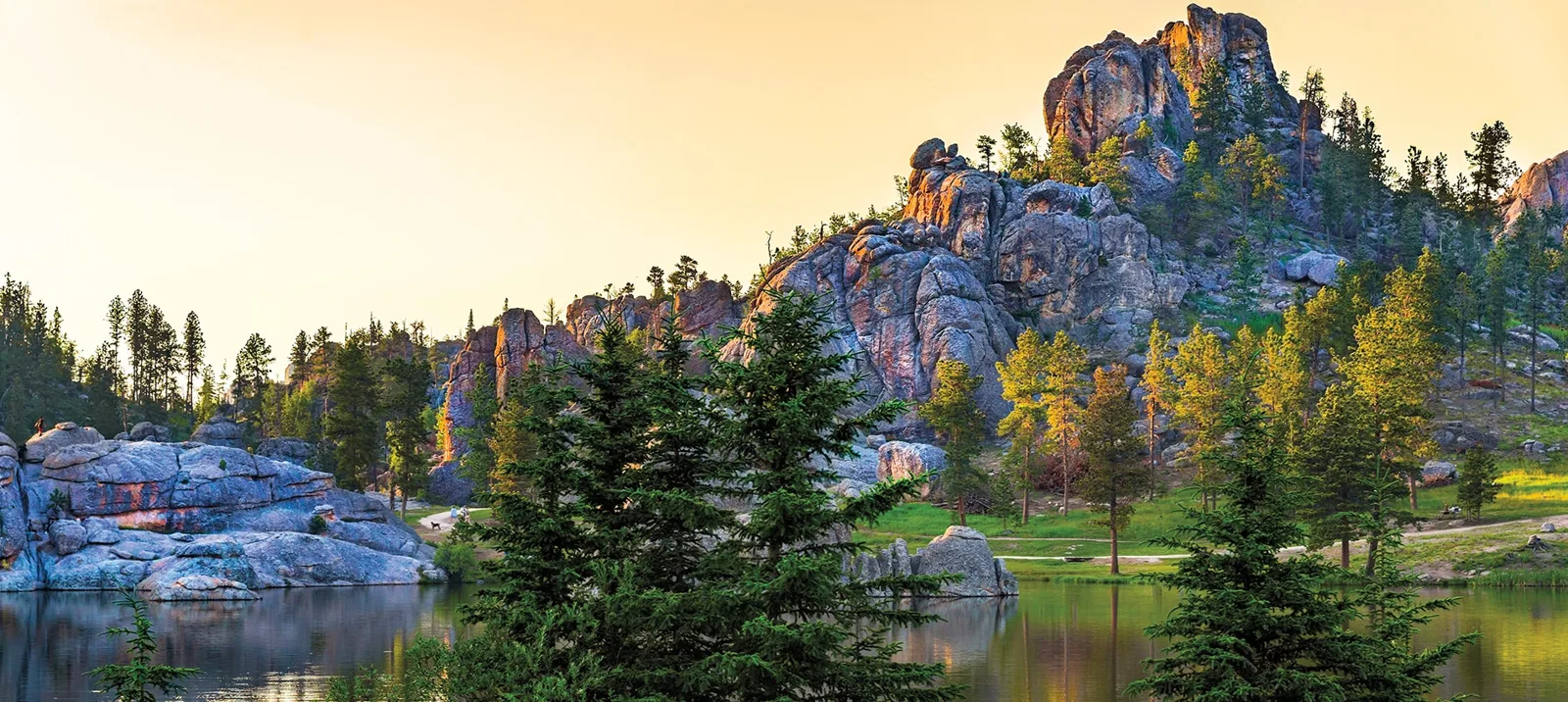 Granite rock spires and pine trees rising above a calm lake in the Black Hills, glowing in warm golden light, with walking paths along the grassy shore.