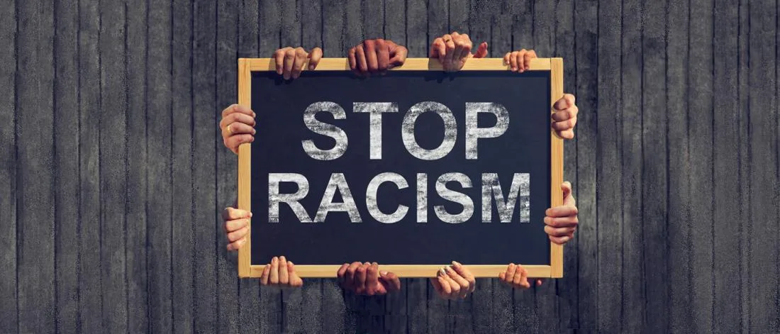 Many hands of different skin tones hold up a wooden-framed chalkboard reading "STOP RACISM" against a dark wooden wall.