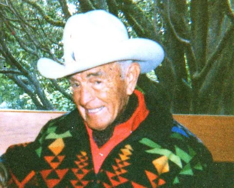 An elderly man in a white cowboy hat smiles, wearing a colorful geometric-patterned blanket jacket over a red shirt, with trees behind him.