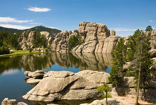 A calm lake reflects towering granite rock formations under a blue sky, with pine trees along the rocky shore in the Black Hills.