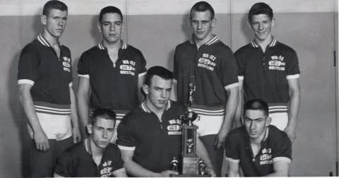 Black-and-white team photo of seven young wrestlers in matching Wa-Hi Wrestling shirts, four standing and three kneeling around a tall trophy.