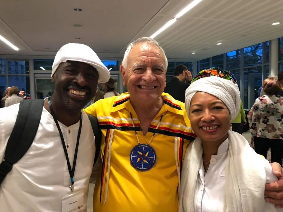 Three smiling people pose together indoors: a man in a white cap and white shirt, an elder in a yellow ribbon shirt with a beaded star medallion, and a woman in a white head wrap and shawl.