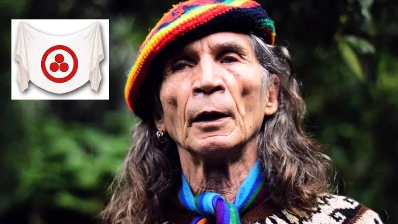 Brother Coyote Alberto Ruz Buenfil, an elder with long gray hair, a rainbow knit cap and a striped scarf, speaking outdoors. An inset shows a white banner with a red circle around three red dots.