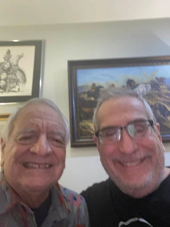 Two smiling older men take a close-up selfie indoors, one in a colorful patterned shirt and one in glasses and a black t-shirt, with framed artwork on the wall behind them.
