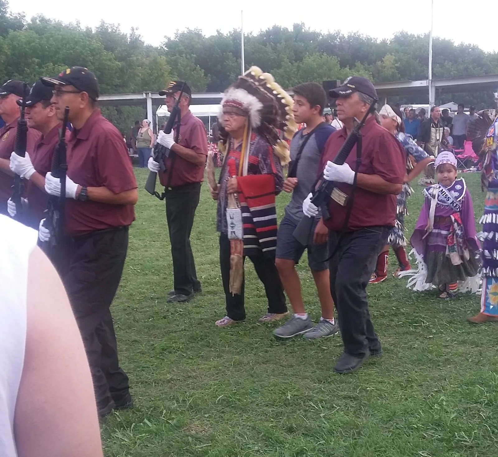 An elder in a feather headdress and a teenage boy walk across a powwow ground, escorted by veterans in maroon shirts and white gloves carrying rifles.
