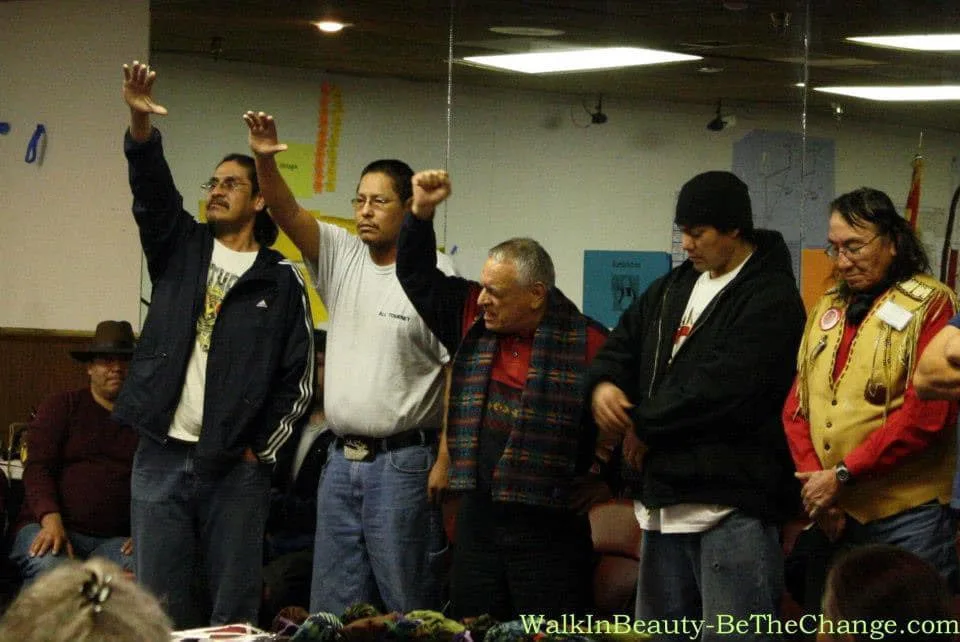 A row of Native men stand indoors in a meeting room, several raising their hands or fists in prayer and solidarity while others bow their heads; an elder wears a beaded buckskin vest.