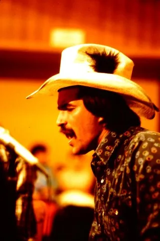 A man with a mustache wearing a light cowboy hat with a dark feather tuft and a patterned shirt, seen in profile against a warm orange background.