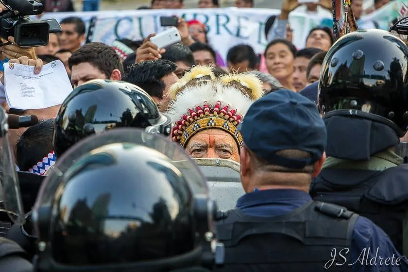 An elder in a beaded and feathered headdress stands face to face with helmeted riot police holding shields, with a crowd and a banner behind him.
