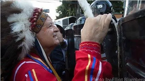 A smiling elder in a feathered headdress and red ribbon shirt rests his hand on a riot police shield as helmeted officers stand close by.