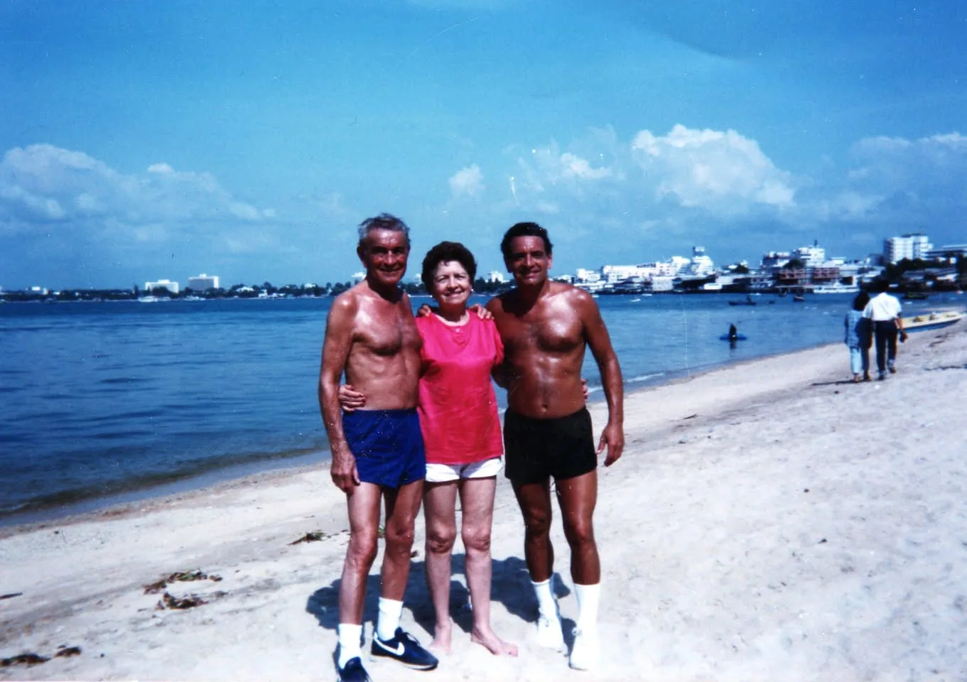 Two shirtless men in shorts and white socks stand on a sunny beach with an older woman in a pink top and white shorts between them, arms around each other, with blue water and a city skyline behind.