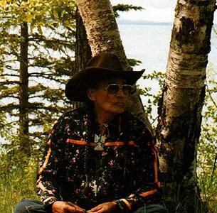 Ernest Tootoosis, wearing a brown cowboy hat, tinted glasses and a dark floral shirt with orange trim, sits between birch trees with a lake behind him.