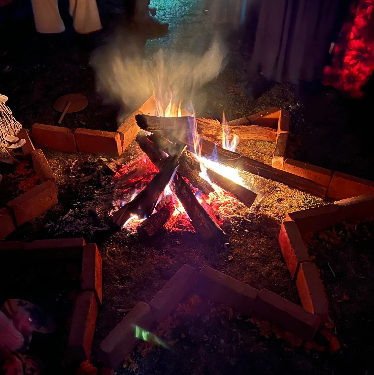 A wood fire burns at night inside a ring of bricks arranged like a star, with smoke rising and people in long garments standing around it.