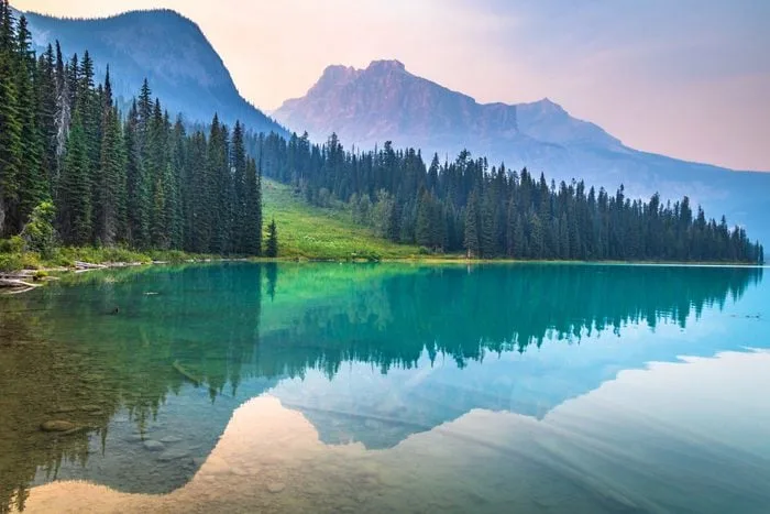 A still turquoise lake mirrors a forest of evergreens and hazy mountain peaks under a soft pink sky.