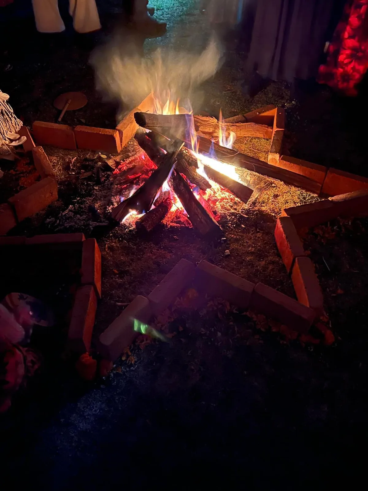 A ceremonial fire burns at night inside a star-shaped border of bricks, with people in ceremonial clothing standing around it.