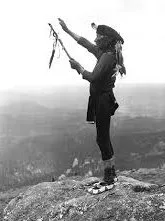 Black-and-white photograph of a Native American man in traditional dress standing on a rocky summit, one arm raised to the sky and holding a feathered staff, with misty hills beyond.