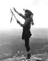 Fotografía en blanco y negro de un hombre indígena de pie sobre una cima rocosa, con el brazo derecho alzado al cielo y una pipa sagrada en la mano, con un valle de fondo.