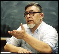 An older man with gray hair, a gray beard and dark-rimmed glasses gestures with one hand as he speaks, wearing a light button-up shirt in front of a chalkboard.