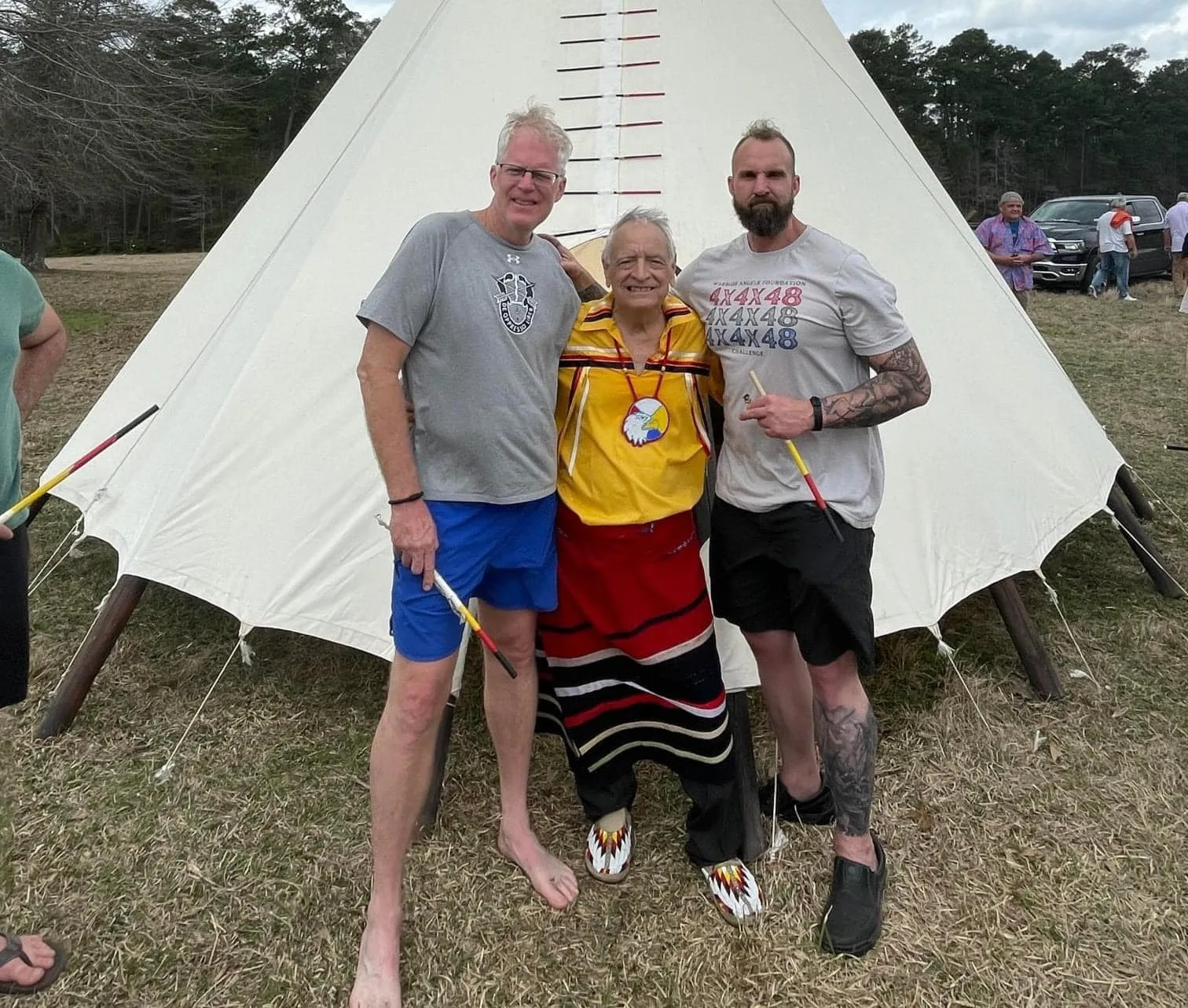Three men stand in front of a white tipi on a grassy field: an elder in a yellow ribbon shirt with an eagle medallion and beaded moccasins, between Chris Miller and Andrew Marr, both holding painted stakes.