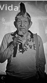 Black-and-white photo of an elderly man in a feathered headband speaking into a microphone, a strap across his chest, with the word "Vida" on a screen behind him.
