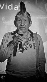 Fotografía en blanco y negro del Gran Anciano Marceliano Jerone, Abuelo Tabaco Verde, con tocado de plumas, hablando por un micrófono.