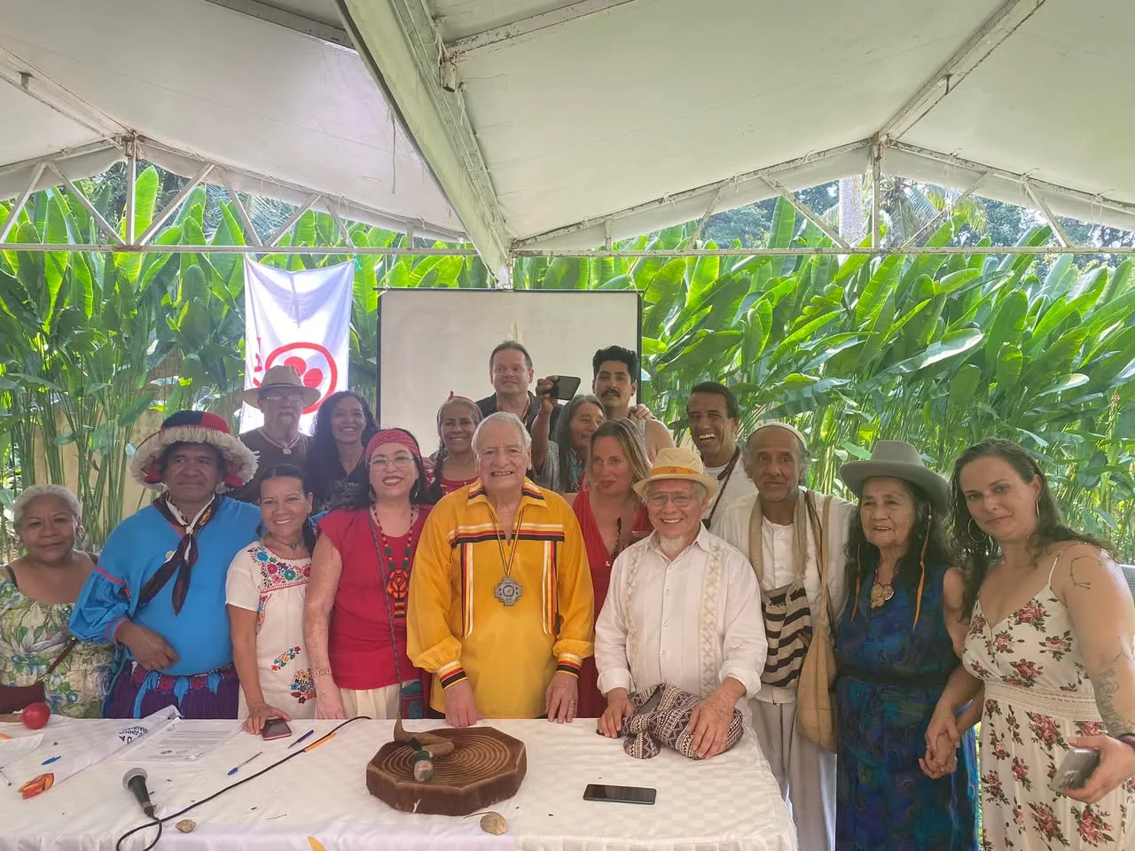 A large group of Indigenous elders and allies pose together under a white tent before tropical greenery, gathered behind a table holding a carved wooden ceremonial piece, papers and a microphone. At center, an elder wears a yellow ribbon shirt and a large medallion.