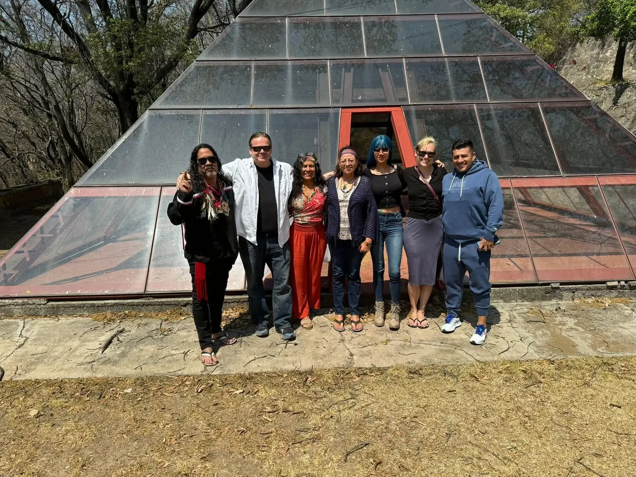Seven people stand arm in arm in sunlight before a large glass pyramid with red frames, one man with long hair wearing a feathered sash and raising his hand.