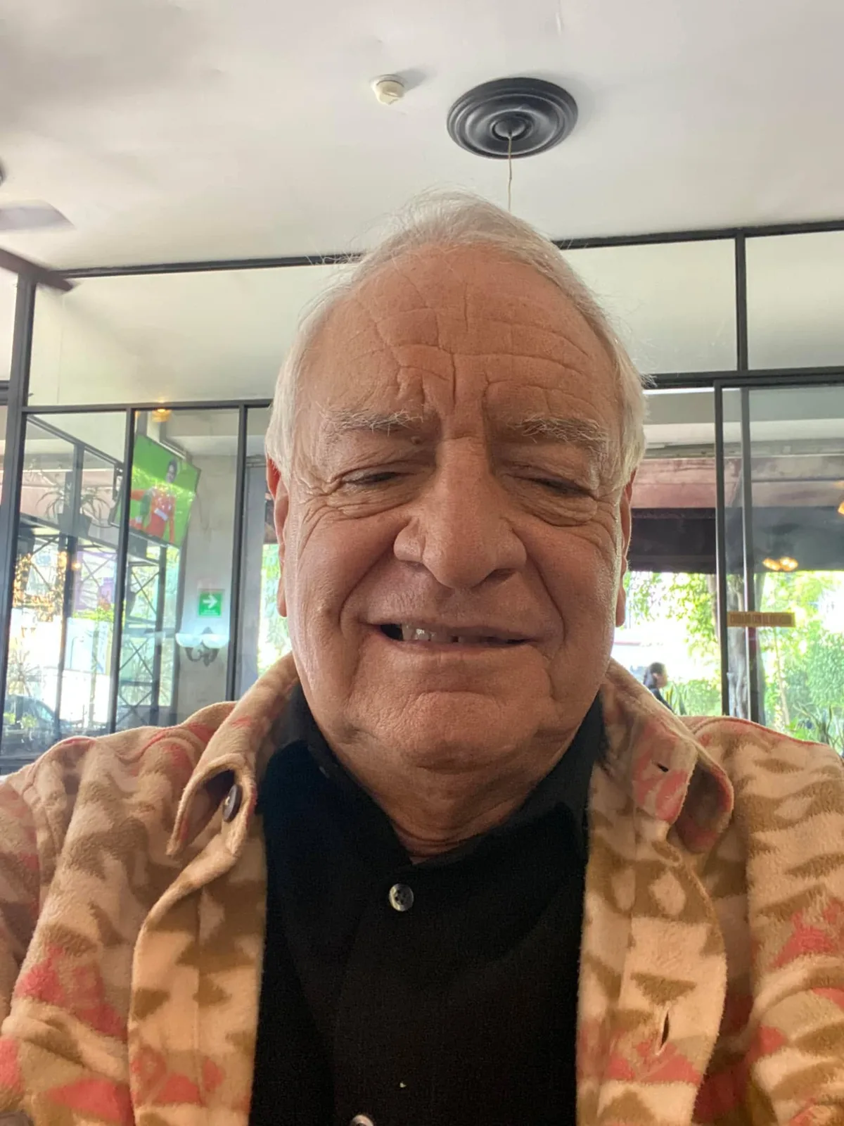 A smiling elderly man with white hair, wearing a patterned pink and tan jacket over a black shirt, takes a selfie inside a restaurant with glass walls.