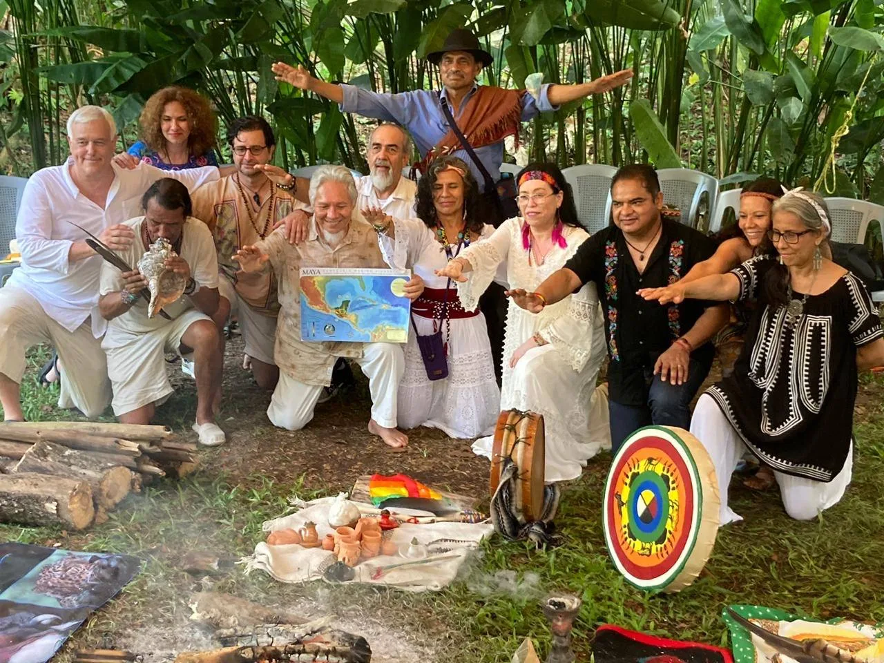 A group in white and embroidered clothing kneels outdoors among tropical plants, arms outstretched toward a ceremonial fire; one man holds a Maya bioregion map, another blows a conch, near a painted drum.