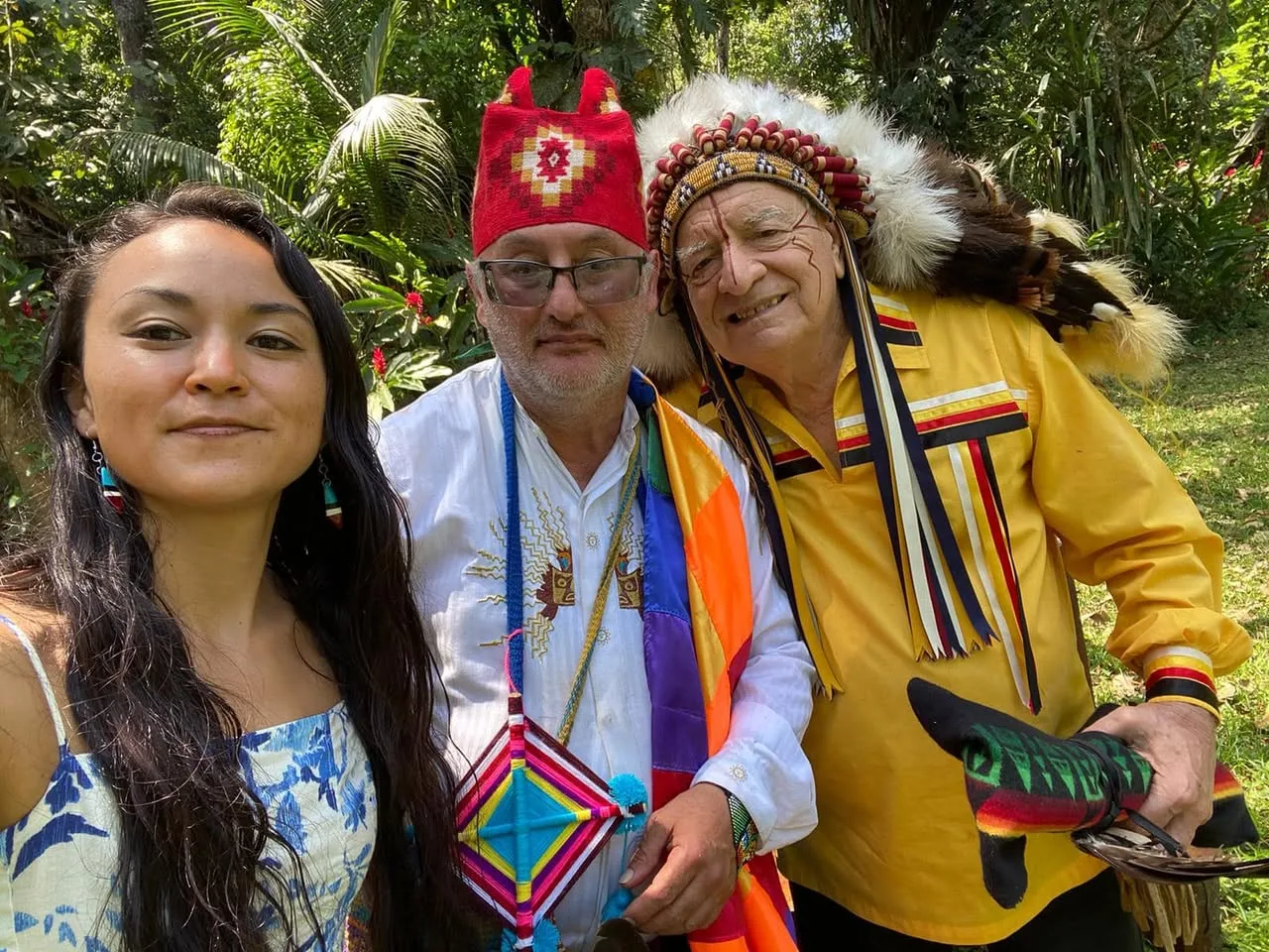 Selfie in a lush tropical garden: Tonja Kumiko in a blue floral dress, Brother Marcelo in a red woven hat and rainbow sash, and Phil Lane Jr. in a feathered headdress and yellow ribbon shirt.