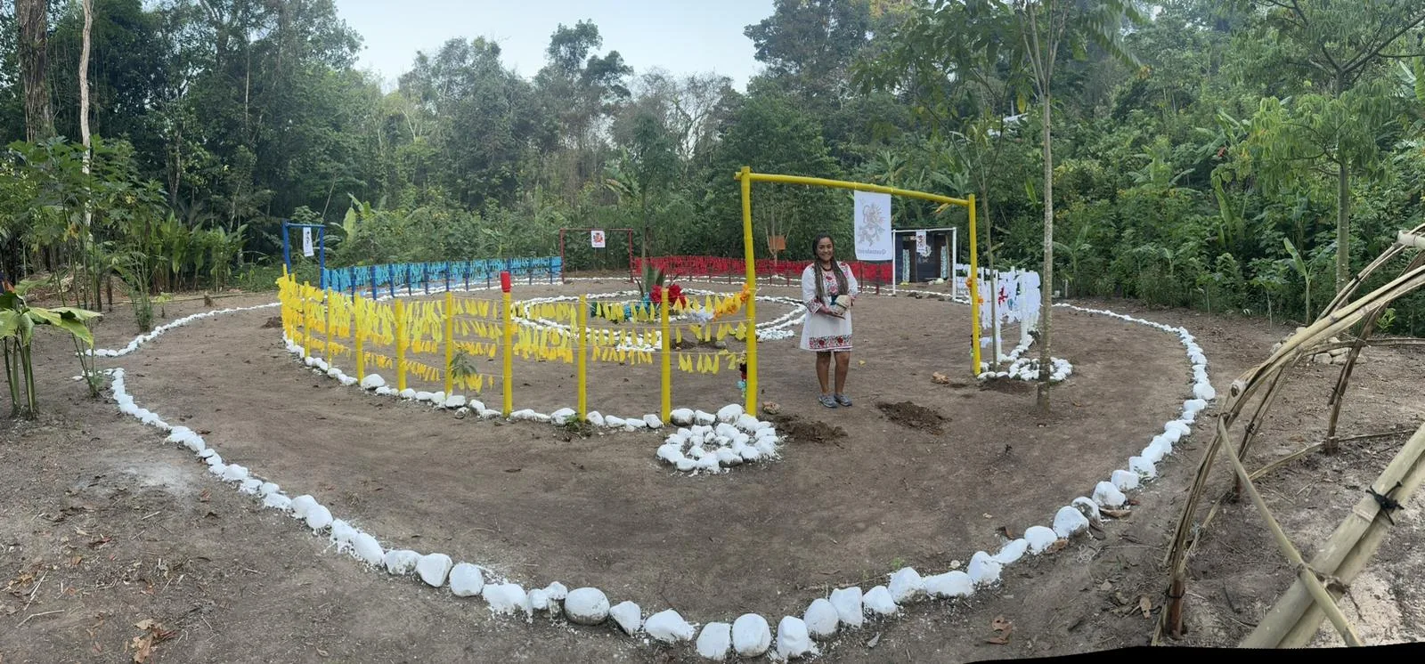 Panoramic view of a ceremonial dance circle outlined with white stones, with yellow, blue, red and white prayer-tie fences; a woman in a white embroidered dress stands inside.