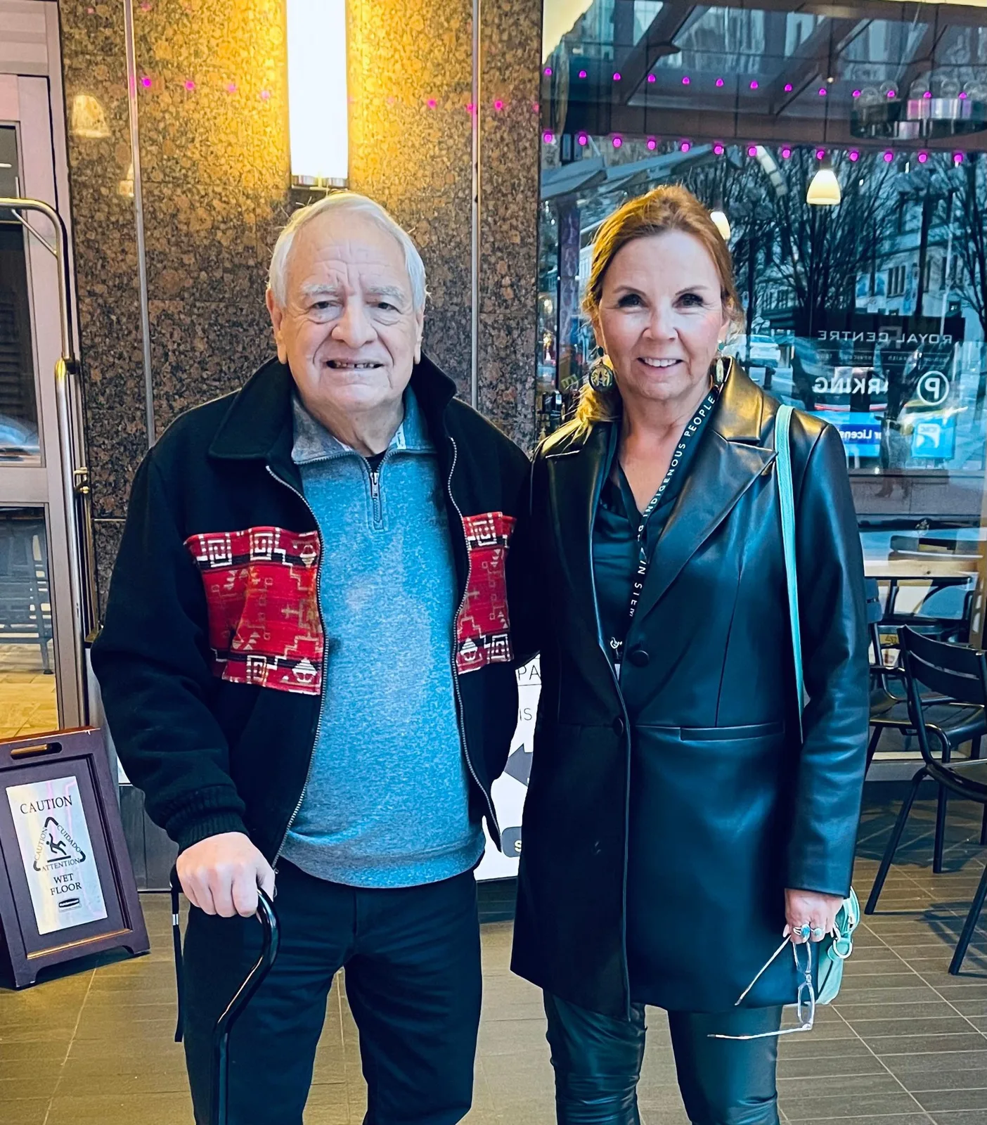 Phil Lane Jr., an elderly man with white hair holding a cane and wearing a black jacket with red patterned panels, stands smiling beside Debbie Beach in a black leather coat in a building lobby.