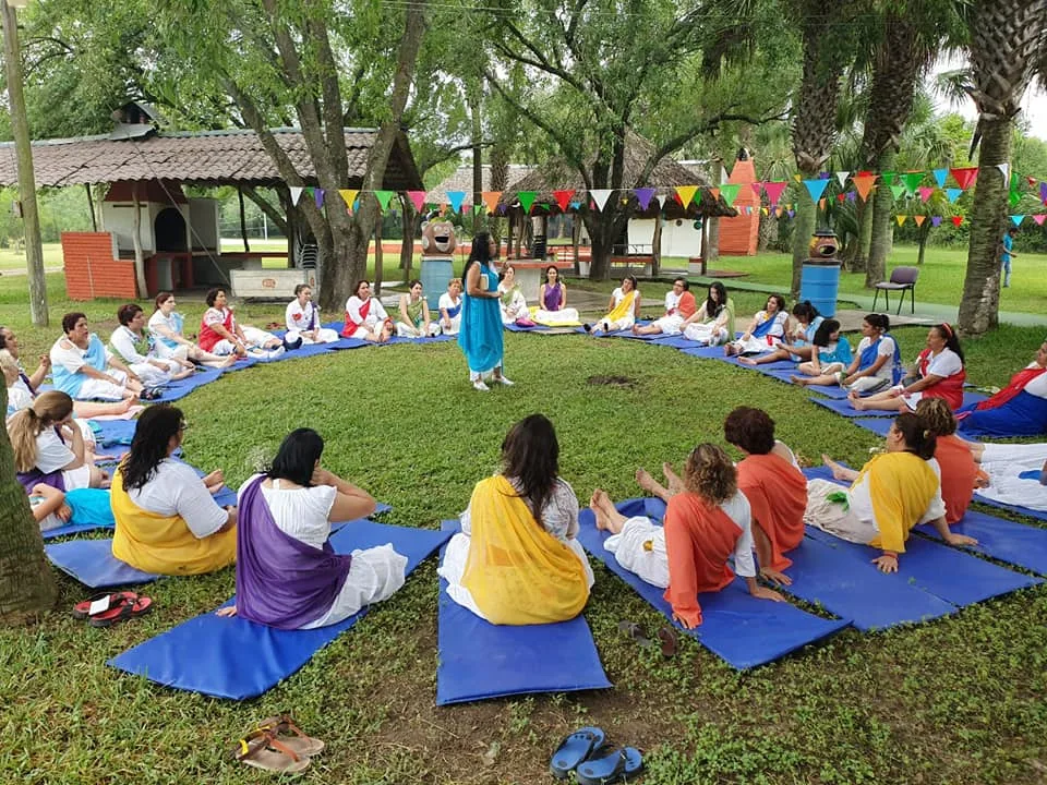 Grupo de mujeres vestidas de blanco con chales de colores, sentadas sobre esteras azules en un gran círculo en el césped, escuchando a una mujer de azul de pie en el centro, bajo árboles y banderines.