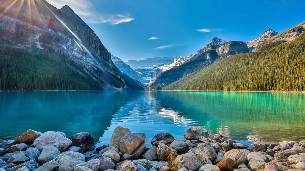 A turquoise mountain lake with a rocky shore in the foreground, framed by pine-forested slopes and snow-capped peaks under a sunlit blue sky.