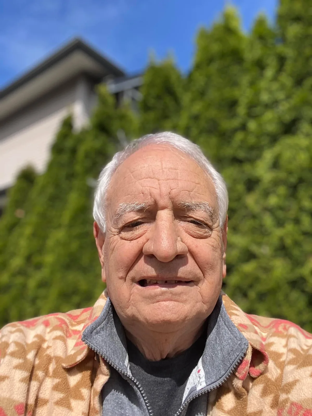 Close-up selfie of Phil Lane Jr., a white-haired elder in a tan patterned fleece jacket, outdoors under blue sky before tall green cedars and a house.