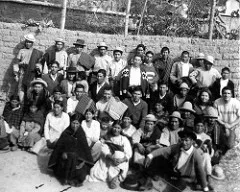 Black-and-white group photo of about thirty Quechua men and women, many in hats and woven blankets, gathered in rows before a stone wall.