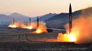 Several missiles launch at once from a flat, open field, trailing bright orange fire and smoke, with hazy mountains in the background.