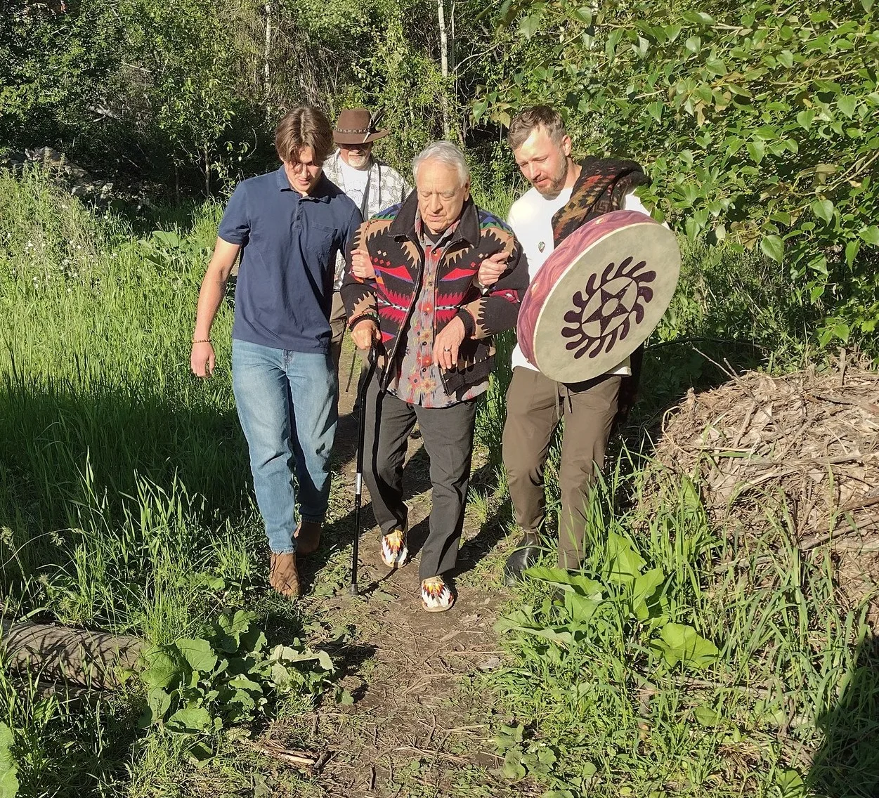An elder with white hair, wearing a colourful patterned jacket and beaded moccasins and using a cane, walks arm in arm with two younger men along a grassy forest path; one carries a hand drum and a man in a hat follows.