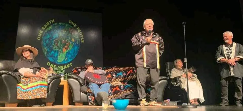 An elder in a ribbon shirt speaks into a microphone on a stage, with four people seated or standing nearby, before a banner reading "One Earth, One People, One Heart" around a painted globe.