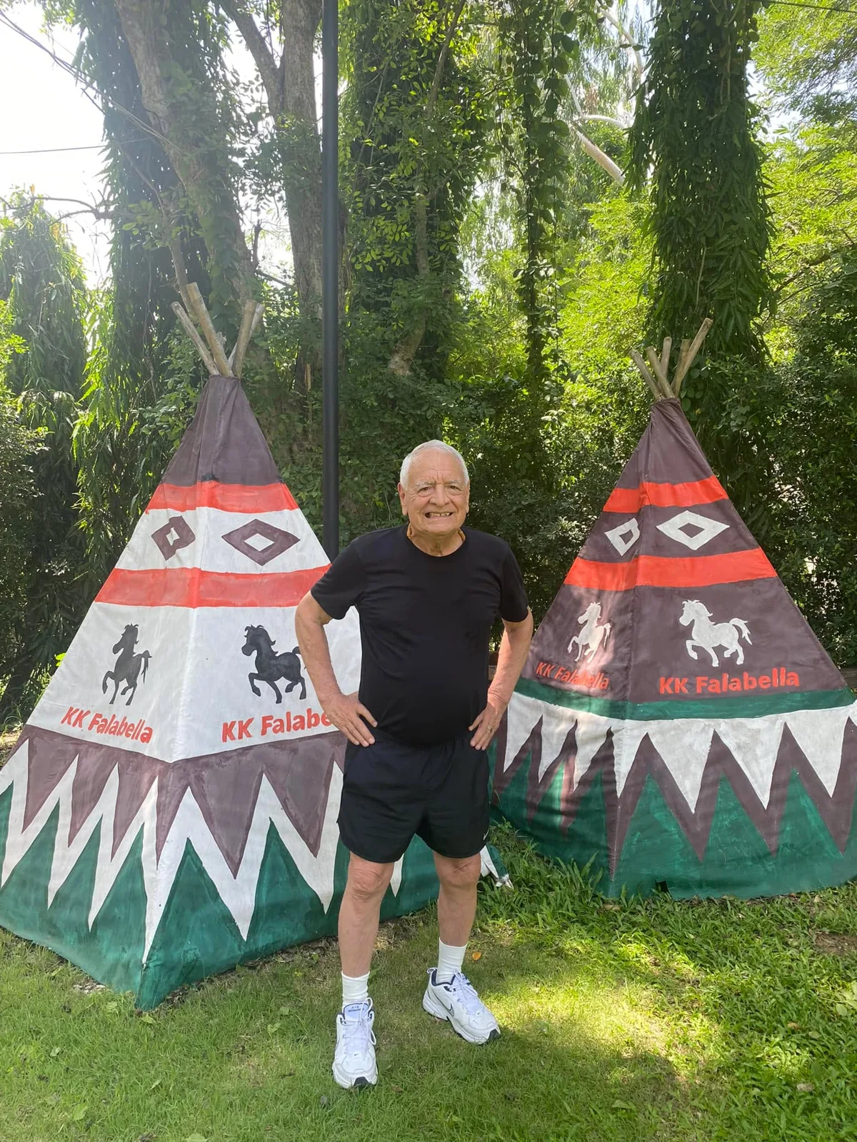 A smiling white-haired elder in a black T-shirt, shorts and sneakers stands with hands on hips on grass between two painted tipis decorated with horses and zigzag patterns, trees behind.
